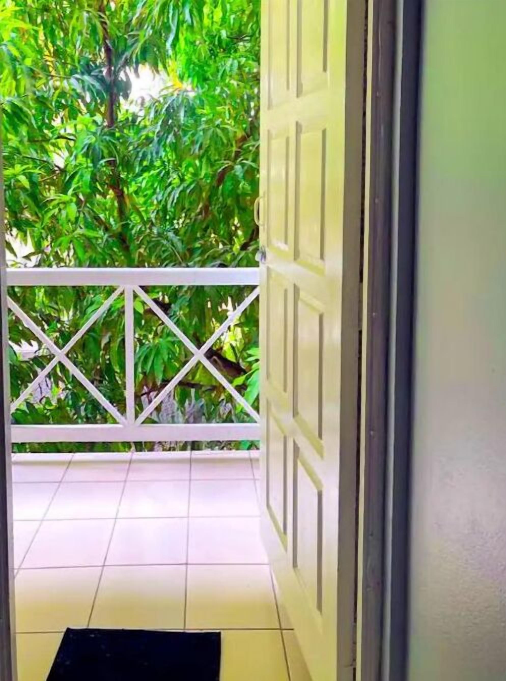 Une porte ouverte donnant sur un balcon doté d’une balustrade blanche et offrant une vue sur un feuillage verdoyant et luxuriant.