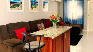 A modern living room with a brown sofa, a small kitchen island, and a vase with flowers. - Gated 2BR Apartment + Enjoy A/C & Wi-Fi + From 100/Night (Kingston 10)