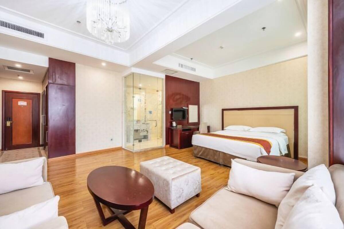 Ein Hotelzimmer mit einem großen Bett, einer Sitzecke mit rundem Tisch, einem Fernseher und einer Glastrennwand.