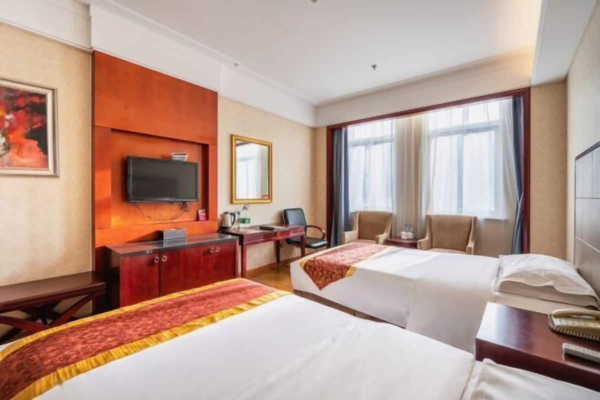 Ein Hotelzimmer mit zwei Betten, einem Schreibtisch, einem Stuhl, einem Fernseher und einem großen Fenster mit Vorhängen.