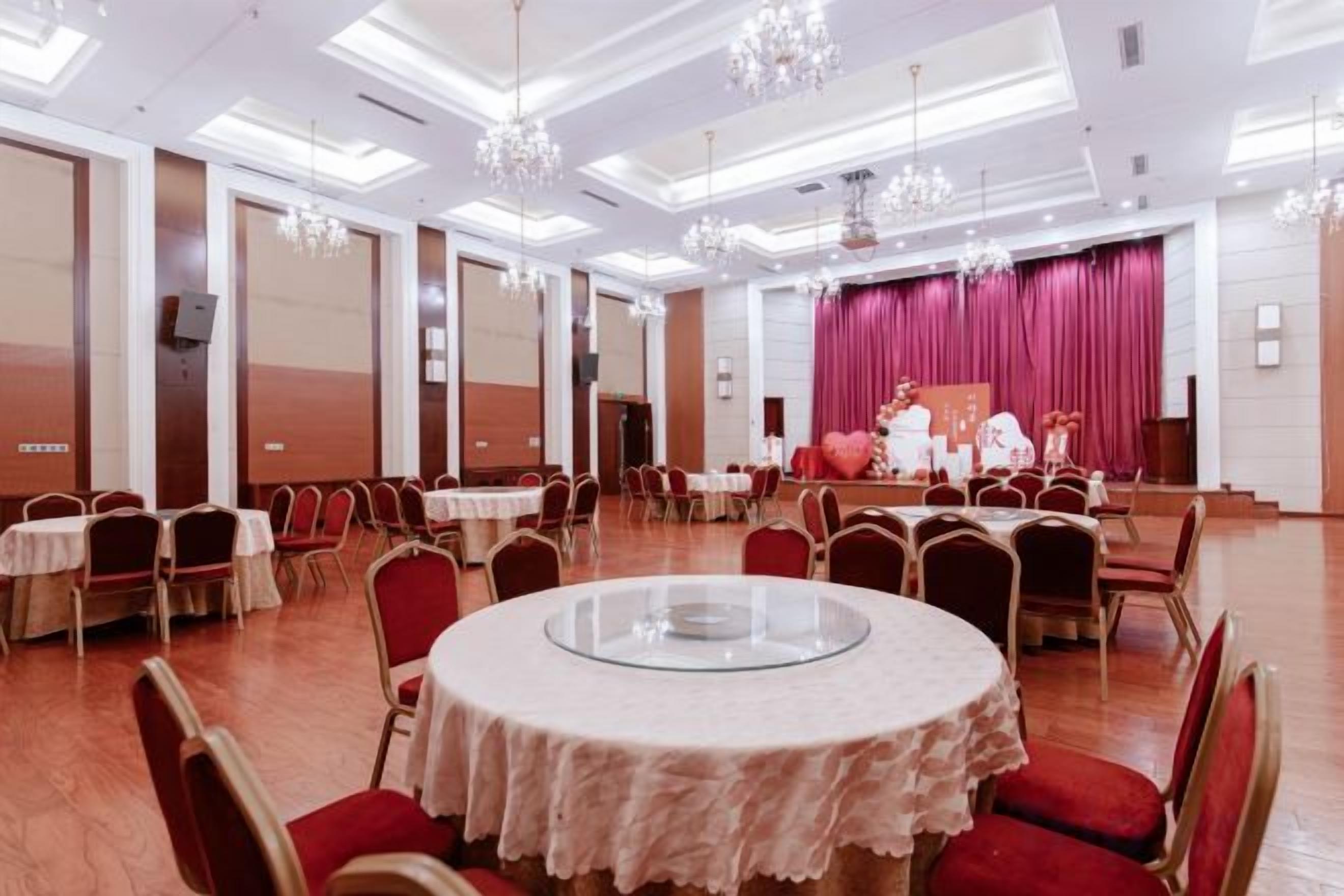 A banquet hall with round tables, chairs, and a stage with a red curtain.
