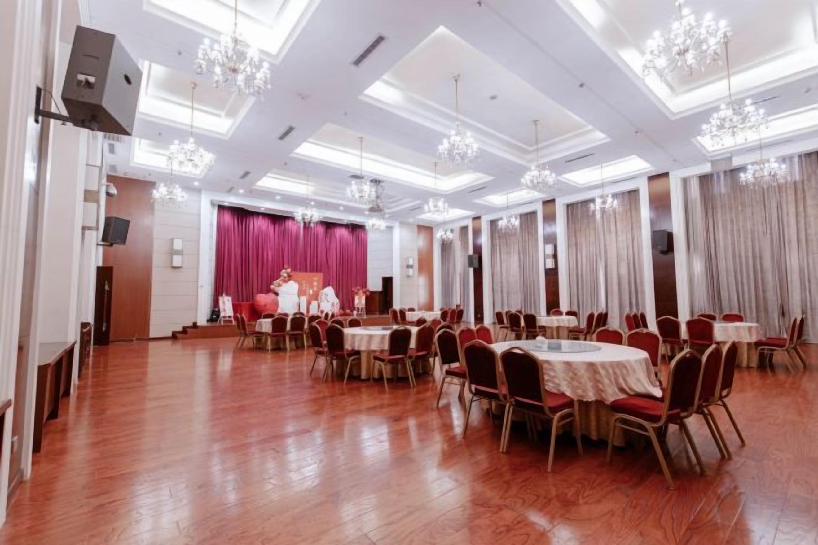 A spacious banquet hall with round tables and chairs, a stage with curtains, and a high ceiling with chandeliers.