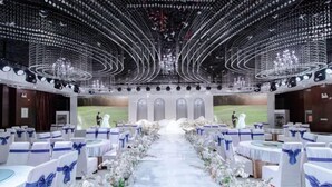 Un salón de bodas con un pasillo central, mesas blancas con cintas azules y un techo decorado con lámparas circulares.