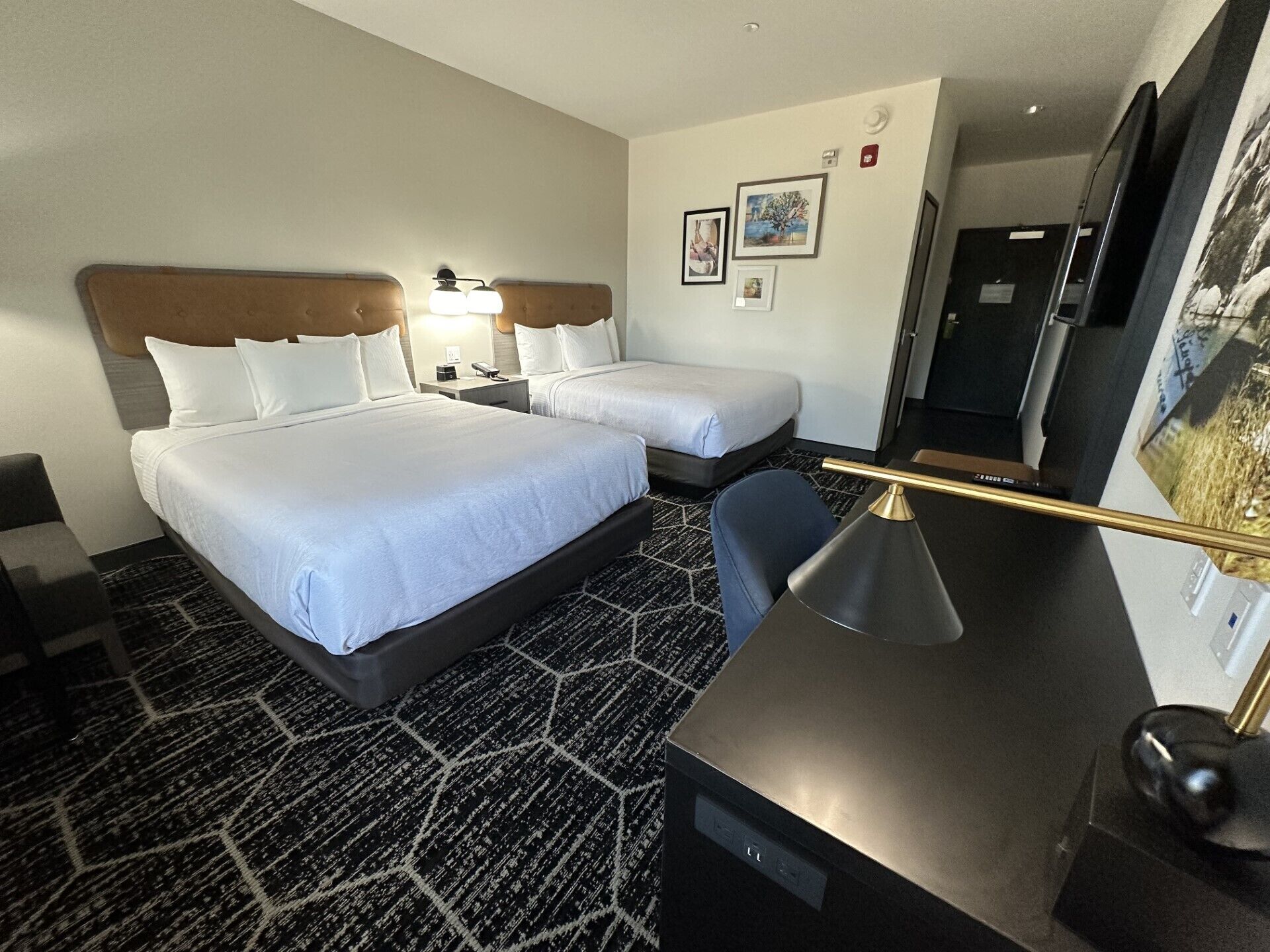 A hotel room with two beds, a desk, a chair, a lamp, and a TV.