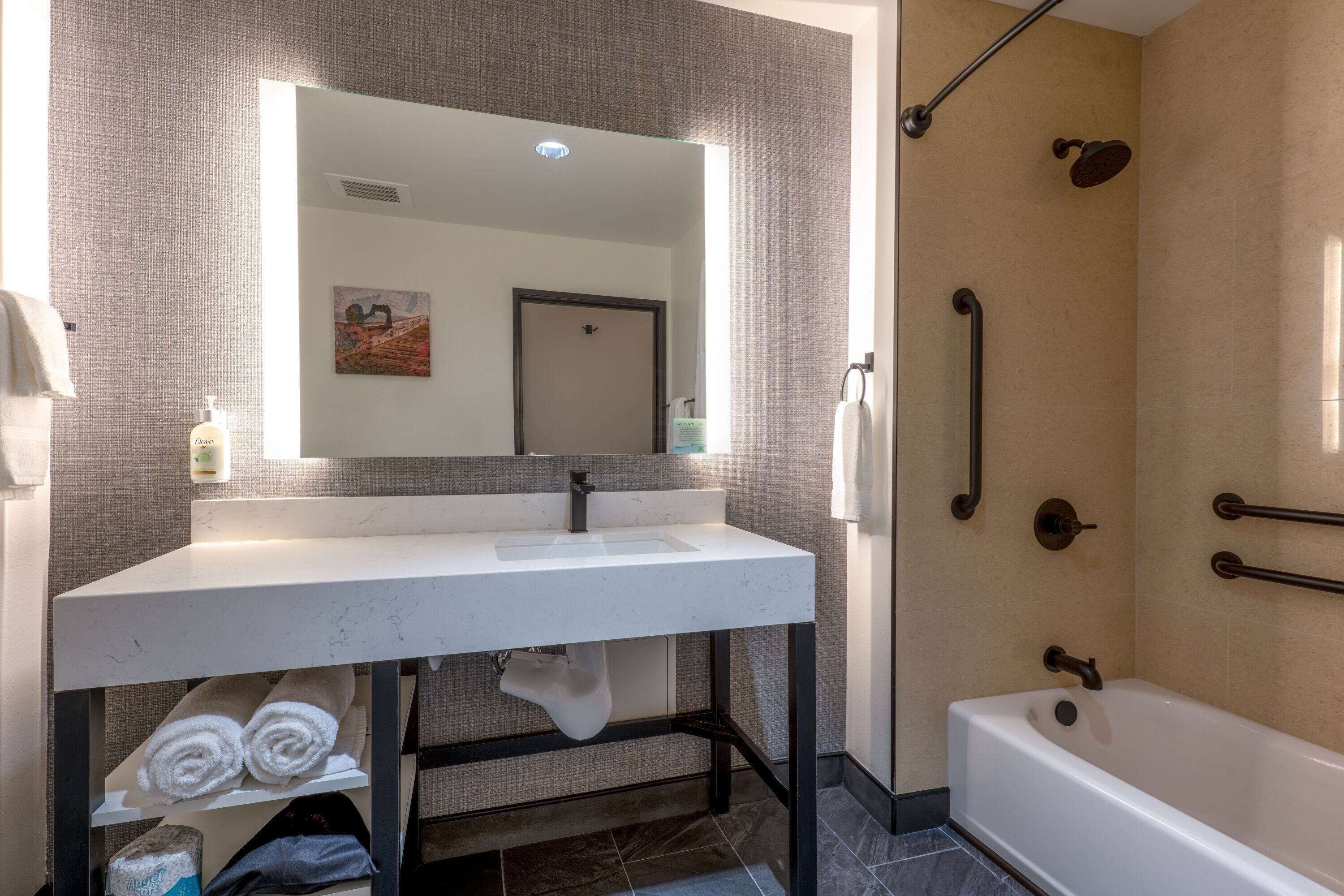 Un baño con un lavabo blanco, un espejo, ducha con barras de apoyo y un perchero para toallas.