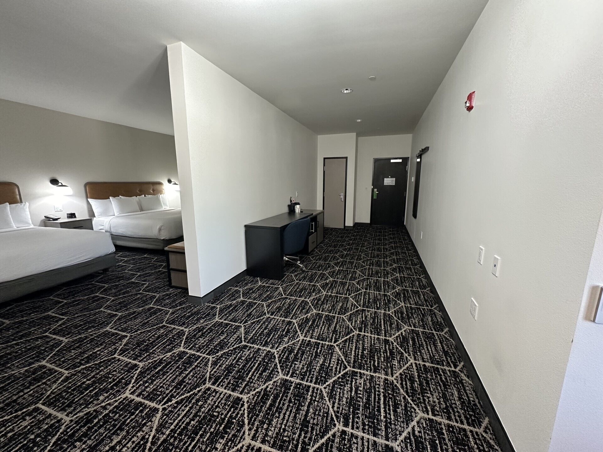 A hotel room with a large bed, a desk, and a chair. There is a door leading to another room.