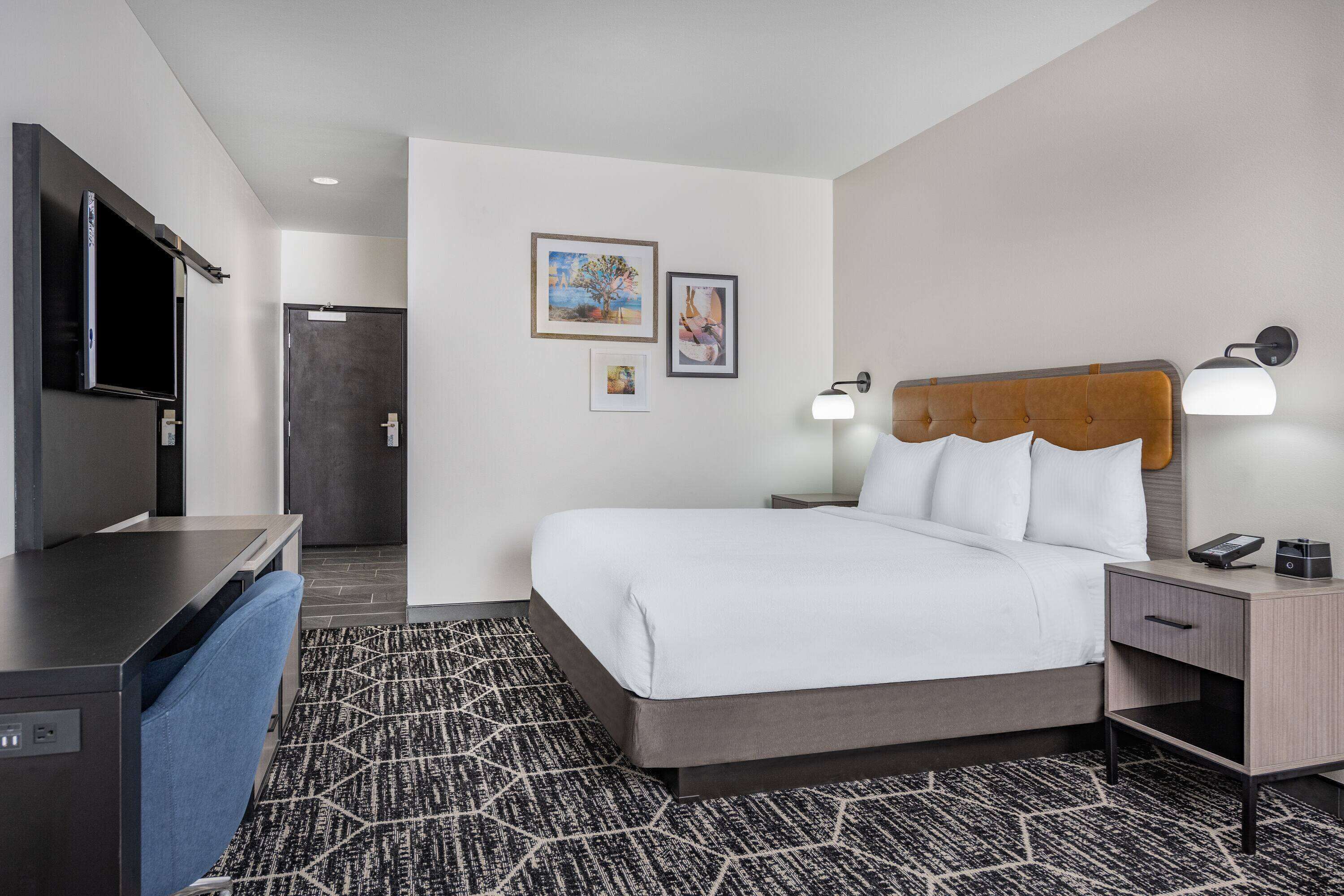 Quarto de hotel com cama, mesas de cabeceira, escrivaninha e televisão.