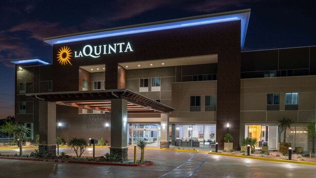A modern hotel with a prominent "La Quinta" sign, featuring a sun logo. The building has multiple floors with large windows and a covered entrance area.