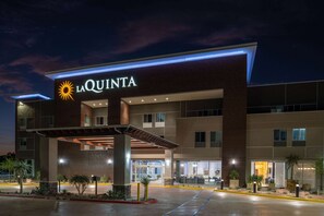 Un hôtel moderne, reconnaissable à son grand panneau « La Quinta », orné d’un logo représentant un soleil. L’immeuble compte plusieurs étages, de grandes fenêtres et une entrée abritée.