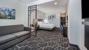 A hotel room with a grey sofa, a bed with white bedding, a desk with a lamp, and a large painting on the wall.