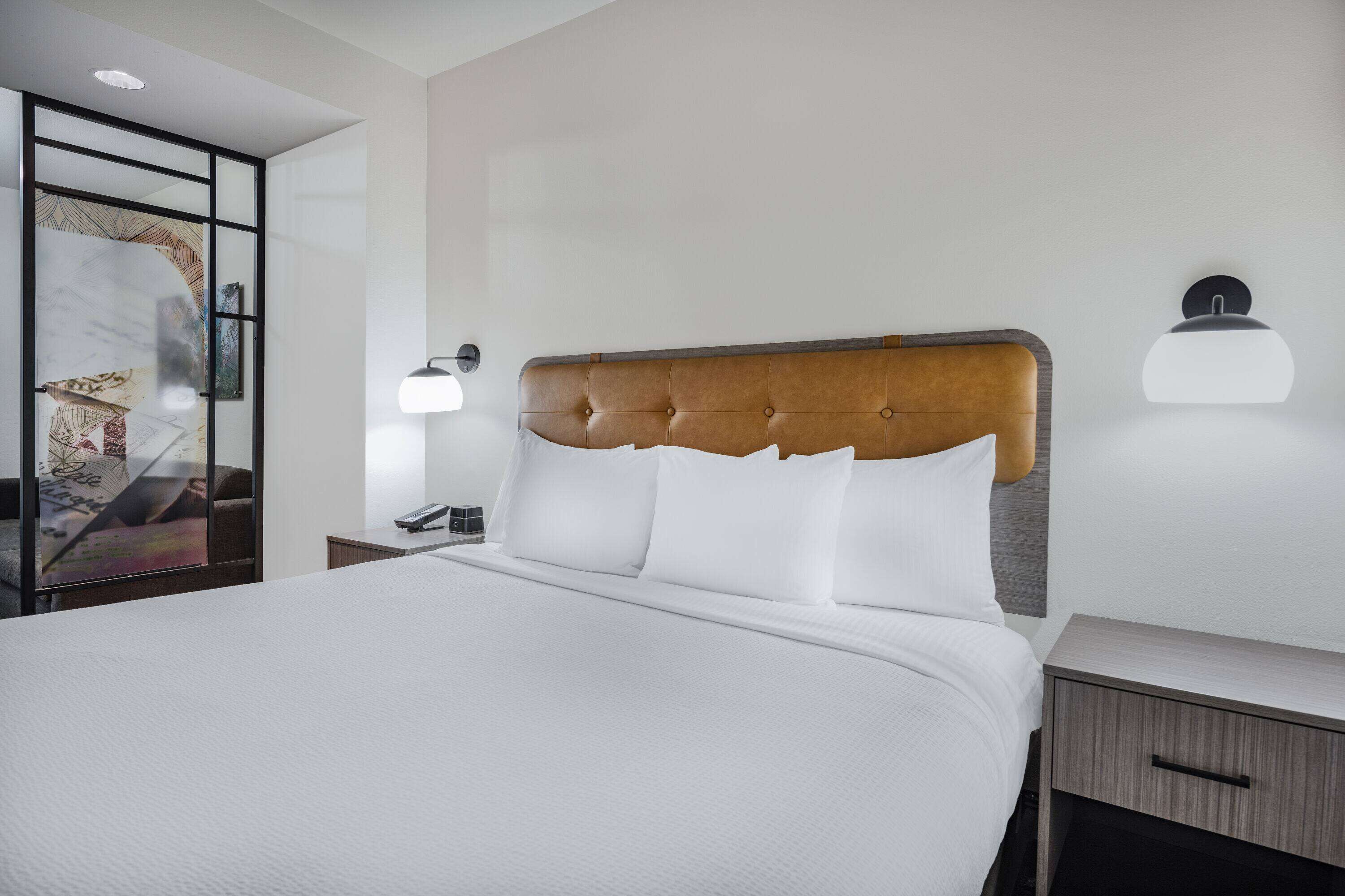 Una cama bien tendida con cabecera capitonada, una mesita de noche con lámpara, y un aplique de pared.