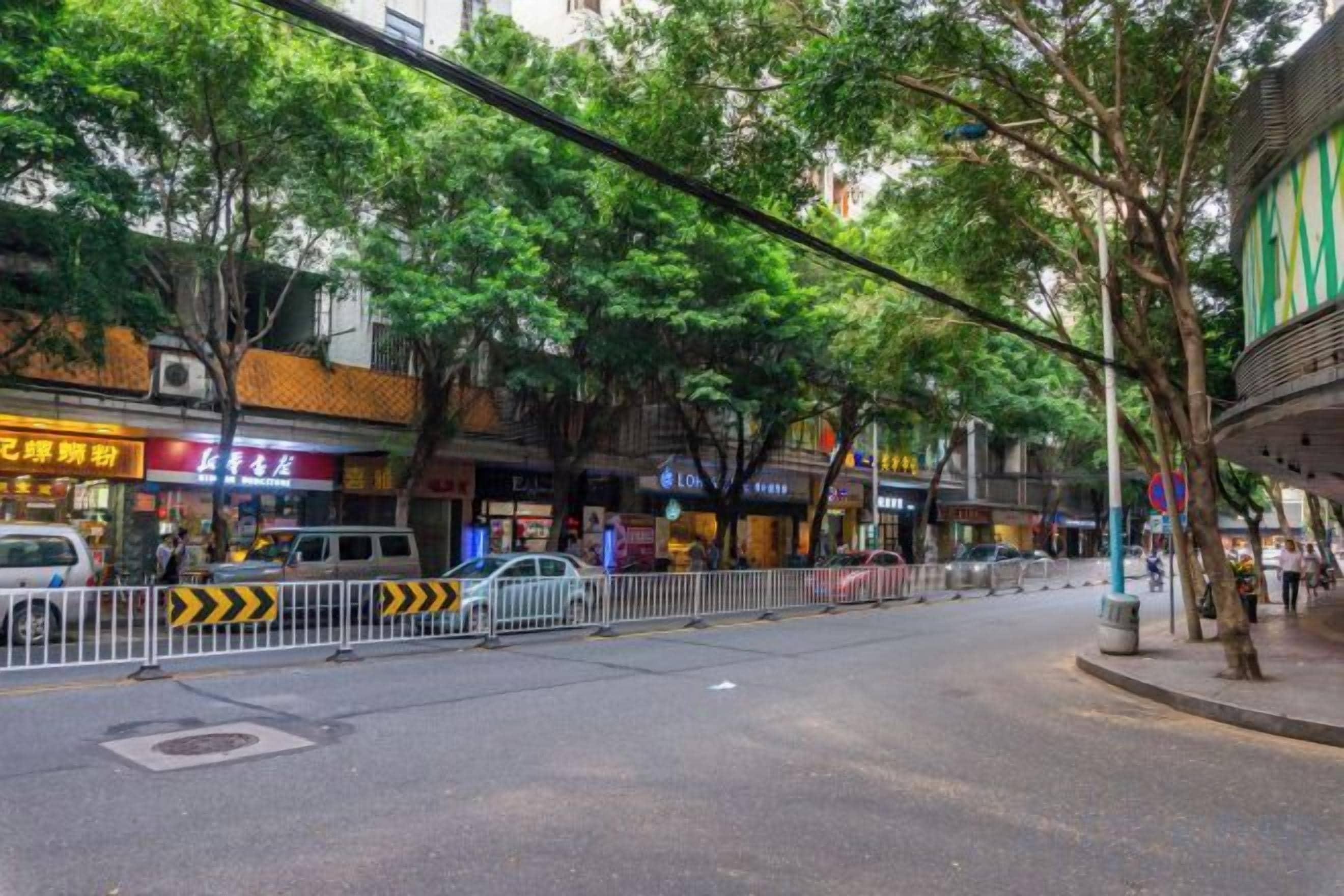 A street with trees, shops, and a fenced-off area.