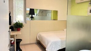 Quarto pequeno e compacto com uma cama de solteiro, mesa de cabeceira com abajur, uma planta em vaso e uma janela.