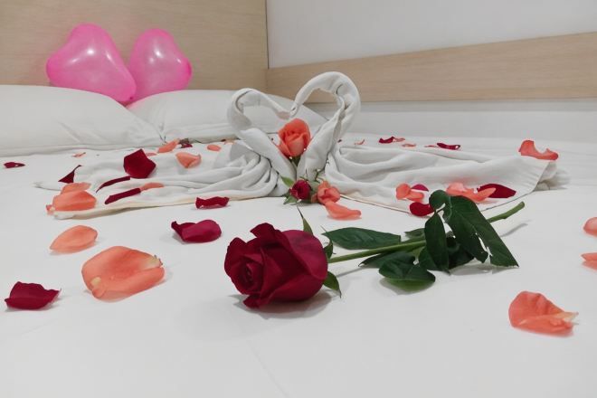 Una cama con pétalos de rosa y una rosa solitaria, almohadas y dos globos rosas.