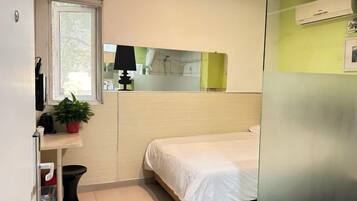 A small, compact hotel room with a single bed, a bedside table with a lamp, a small potted plant, and a window.