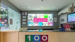 Uma área de recepção com balcão de vidro, um sinal com o número '100', certificados emoldurados e uma parede com painel digital.