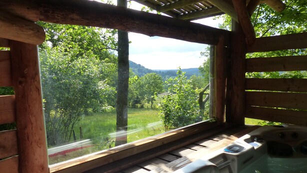 A wooden cabin with a hot tub and a large window overlooking a green landscape.