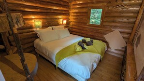 A cozy log cabin room with a bed, two wall-mounted lamps, and a window with a view of trees.