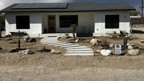 A single-story house with a flat roof, solar panels, a front door, and a small patio area with a table and chairs.