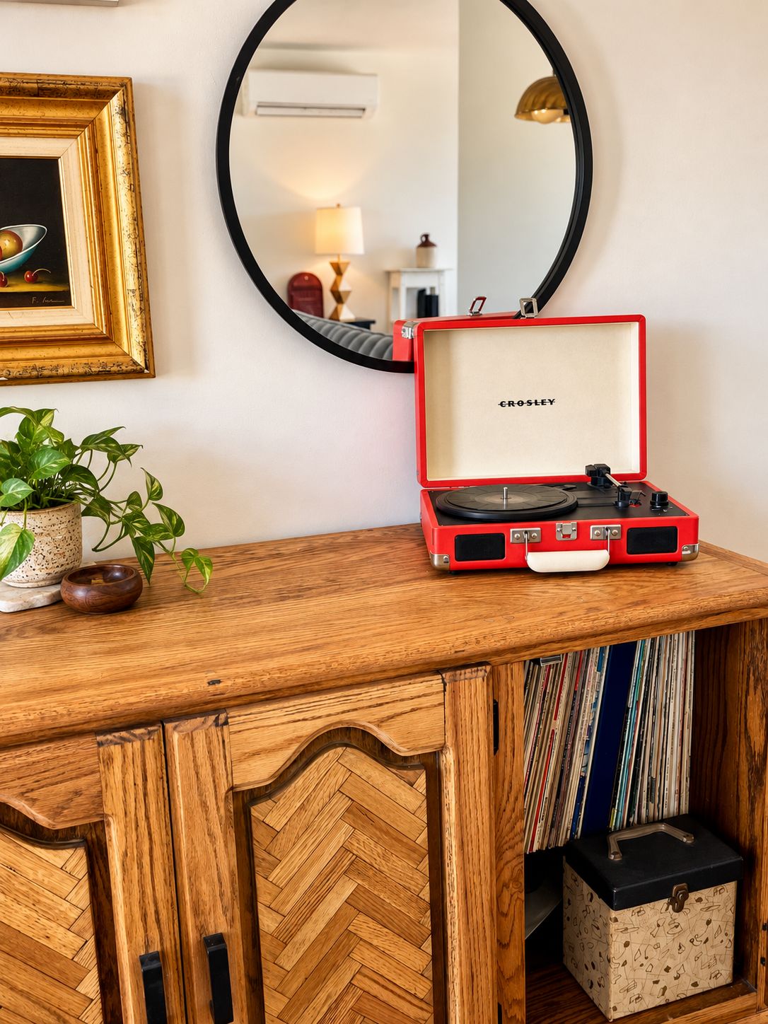Un bureau en bois sur lequel se trouve une valise rouge contenant une platine, un tableau encadré, un miroir et une plante en pot.