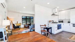 A modern kitchen with white cabinets, a wooden dining table, and a view of the outdoors through large windows.