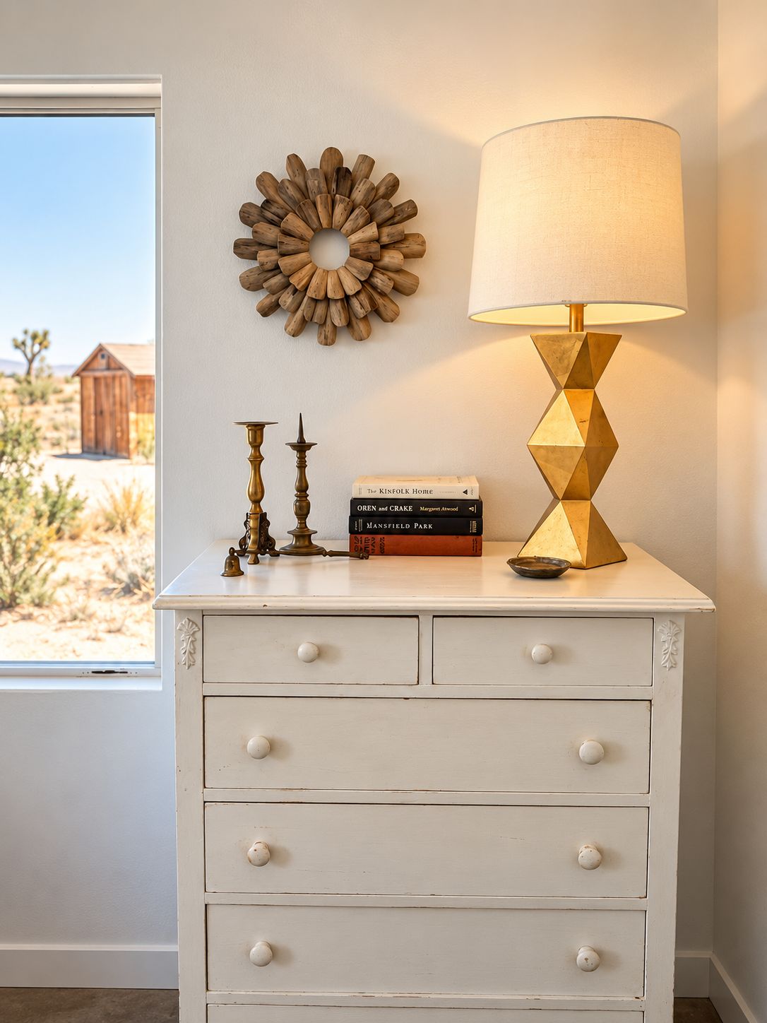 A white dresser with drawers, a wall-mounted decorative sunburst, a table lamp, and a window with a view of a desert landscape.