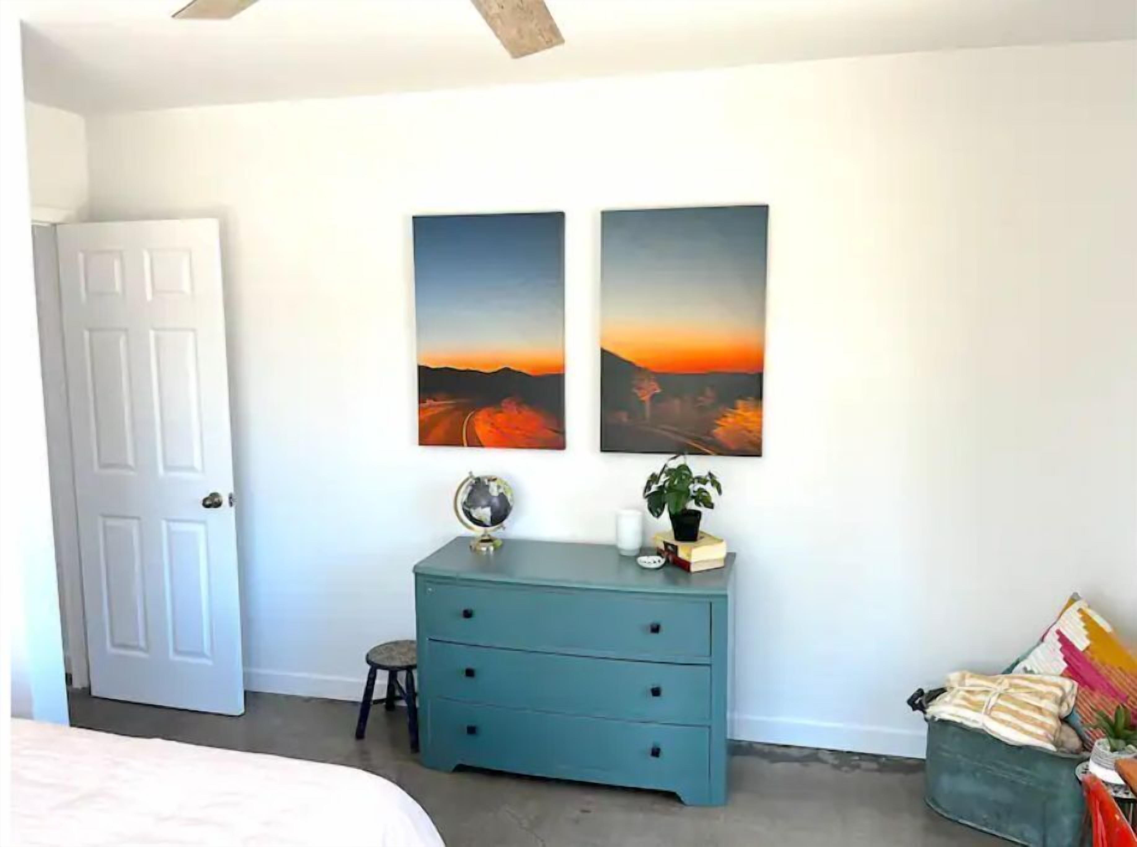 Une chambre avec une porte blanche, une commode turquoise, deux tableaux représentant des paysages, un globe terrestre et un petit tabouret.
