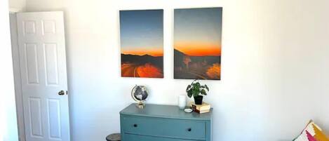 A bedroom with a white door, a teal dresser, two landscape paintings, a globe, and a small stool.