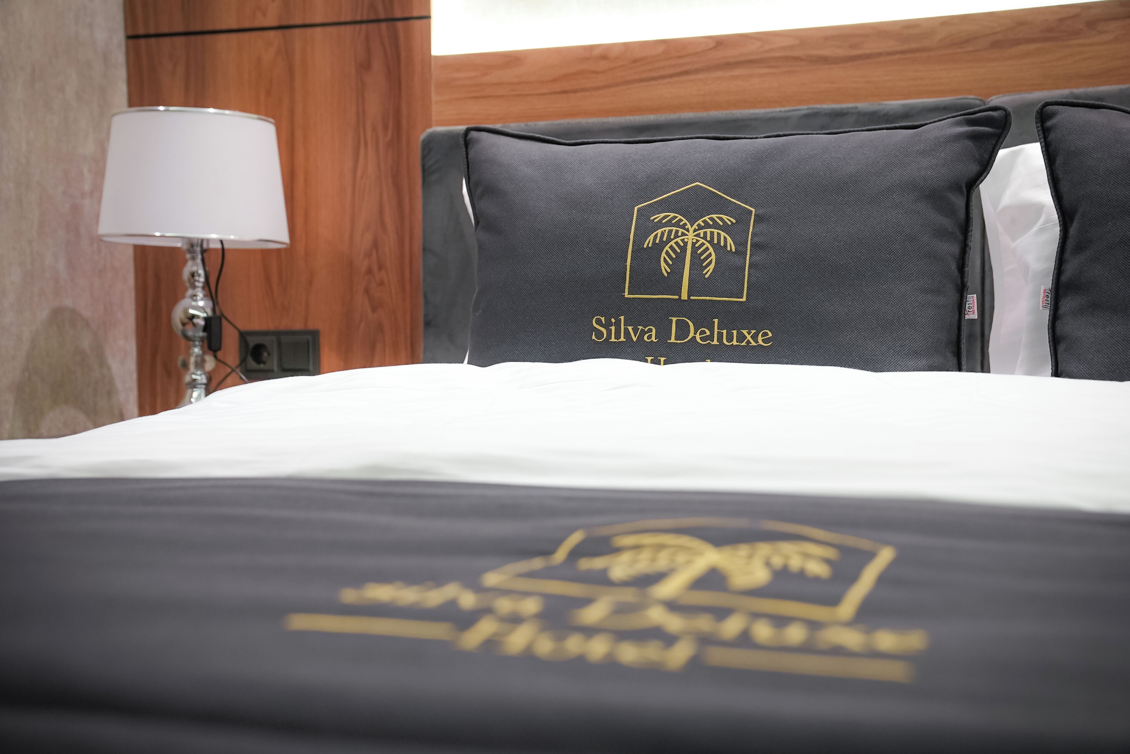 Habitación de hotel con una cama que presenta una almohada negra con una palmera y texto, una colcha blanca y un cabecero de madera.
