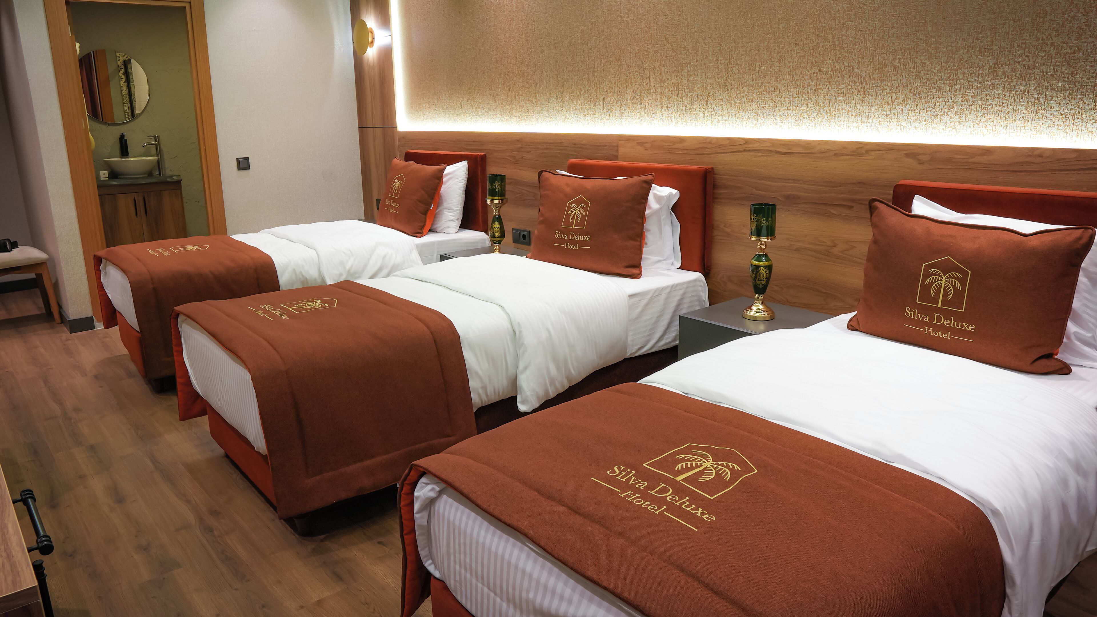 Habitación de hotel con tres camas, cada una con cabecero marrón y ropa de cama blanca. A la derecha, un cabecero de madera, una mesita con una lámpara verde y, a la izquierda, un espejo en la pared.