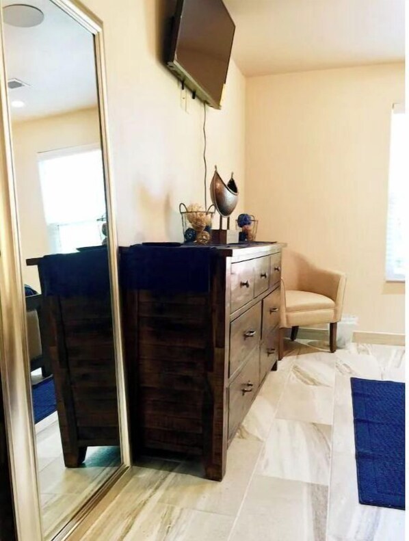 A bedroom with a large mirror, a dresser with drawers, a chair, and a television mounted on the wall.