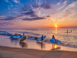 A beach scene with people carrying surfboards, a sunset in the background, and calm ocean waves. - Our Napping House, 5 min from Bethany beach, Sleeps 10, Pool, Pickleball, wifi (Ocean View)