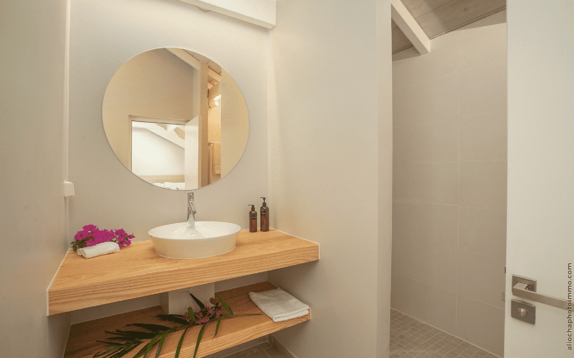 A bathroom with a round mirror, a white sink, wooden shelves, and a shower area.