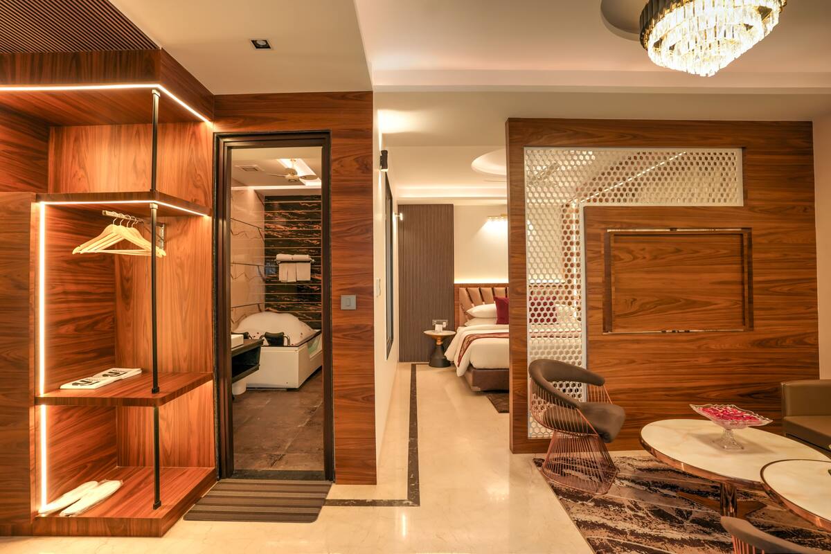 A modern hotel room with a wooden accent wall, a mirror, a bed with a white headboard, a small table with a bowl, and a chandelier.