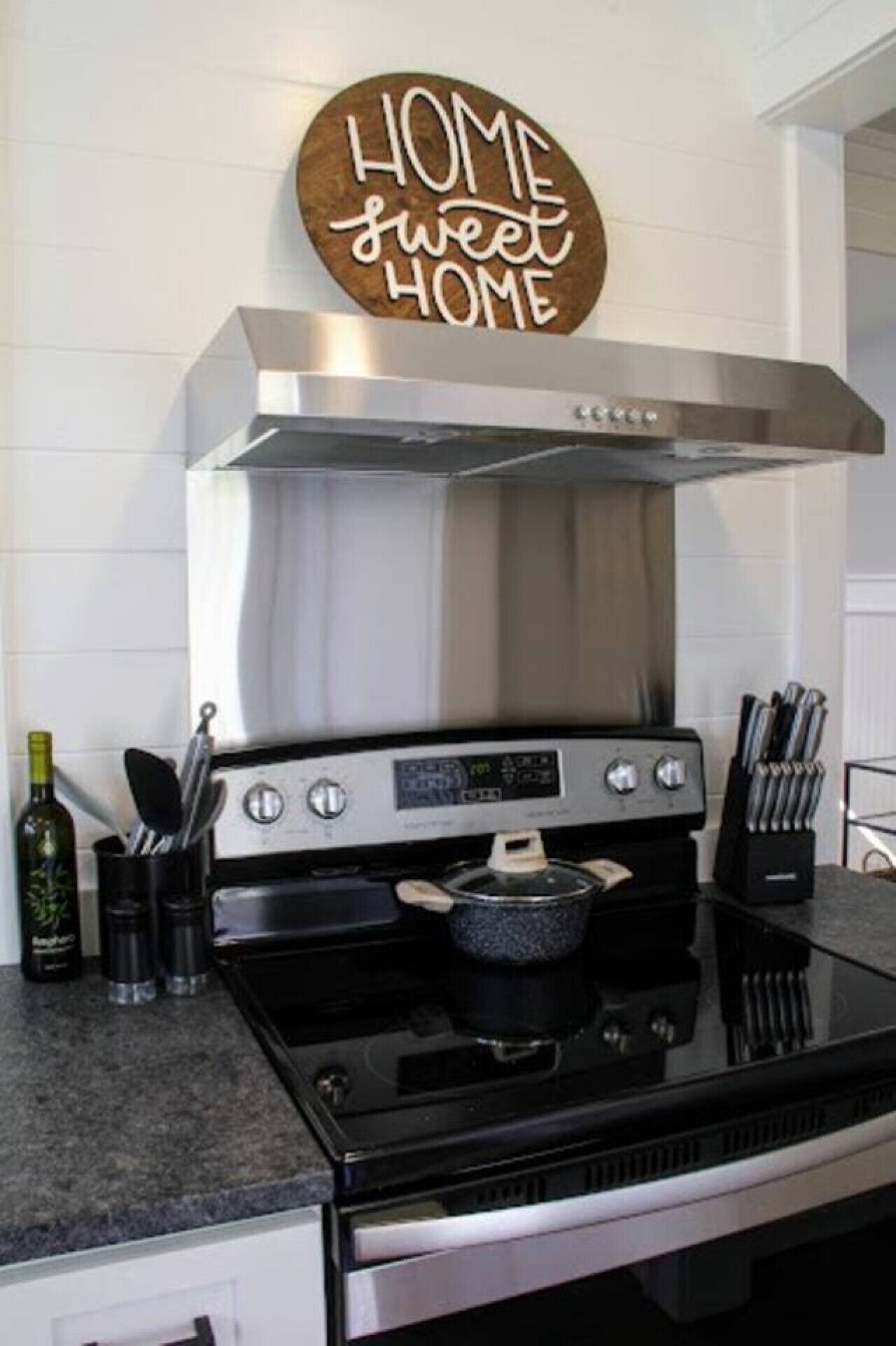 Una cucina con una cappa in acciaio inossidabile, un piano cottura nero e una pentola sul fuoco. Sul bancone c'è un portacoltelli e una bottiglia d'olio d'oliva. Un cartello sopra il piano cottura recita "HOME sweet HOME".