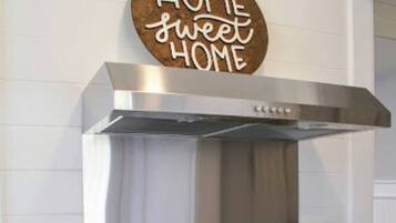 Una cucina con una cappa in acciaio inossidabile, un piano cottura nero e una pentola sul fuoco. Sul bancone c'è un portacoltelli e una bottiglia d'olio d'oliva. Un cartello sopra il piano cottura recita "HOME sweet HOME".