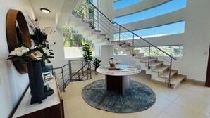 Un interior moderno con una mesa redonda central, una escalera con barandillas de vidrio y vistas al exterior con vegetación.