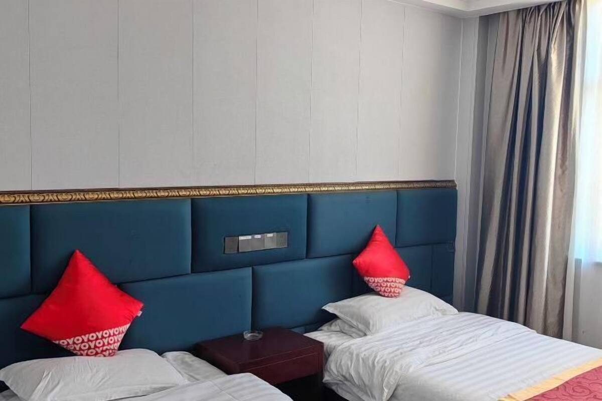 A hotel room with a blue headboard, two beds, red and white pillows, a small wooden nightstand, and a window with curtains.