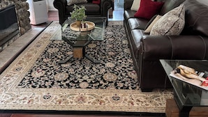 Un salon avec une cheminée en pierre, un canapé en cuir noir, une table basse en verre et un tapis à motifs.