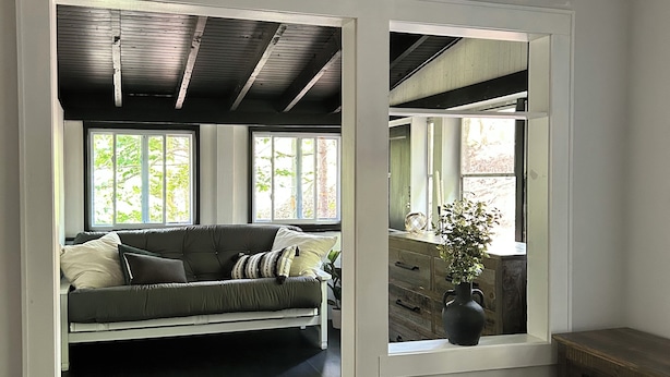 Une pièce avec un plancher en bois, un canapé gris avec des coussins, une commode en bois et une fenêtre donnant sur des arbres.