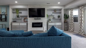 A modern living room with a stone fireplace, a flat-screen TV mounted above it, and a blue sofa with matching cushions. - Beautiful House overlooking Lake Chemung, with a Private Chef Tasting option. (Howell)