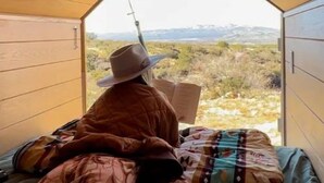 Een persoon zit op een bed in een tentachtige kamer met uitzicht op een landschap.
