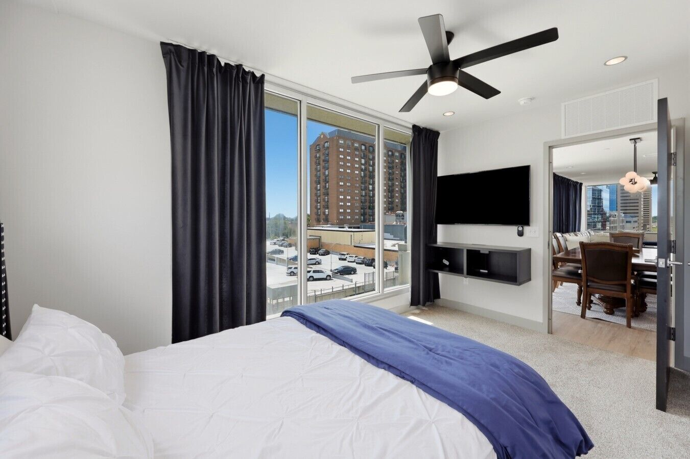 A hotel room with a bed, a ceiling fan, a TV, a desk with a chair, and a view of the cityscape.