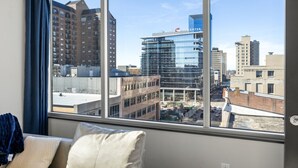 A hotel room with a large window offering a view of a cityscape, including a modern building with a glass facade and a traditional brick building. - Penthouse @ 175 LEX -Walk to Rupp Arena on Main St (Lexington)