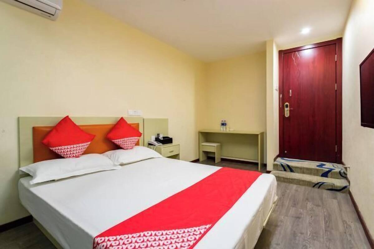 A hotel room with a bed, a desk, a chair, a wardrobe, and an air conditioning unit.
