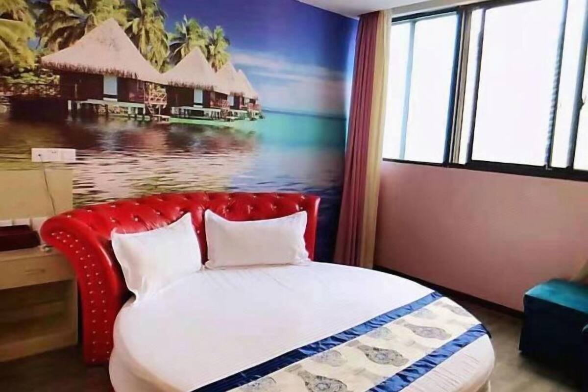 A hotel room with a bed, a red upholstered headboard, a window with curtains, and a mural of a tropical beach scene.