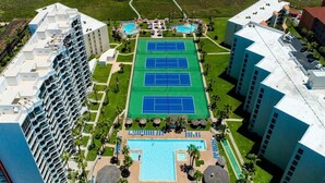 Un hotel con varios pisos, cancha de tenis, piscina y zona de playa.