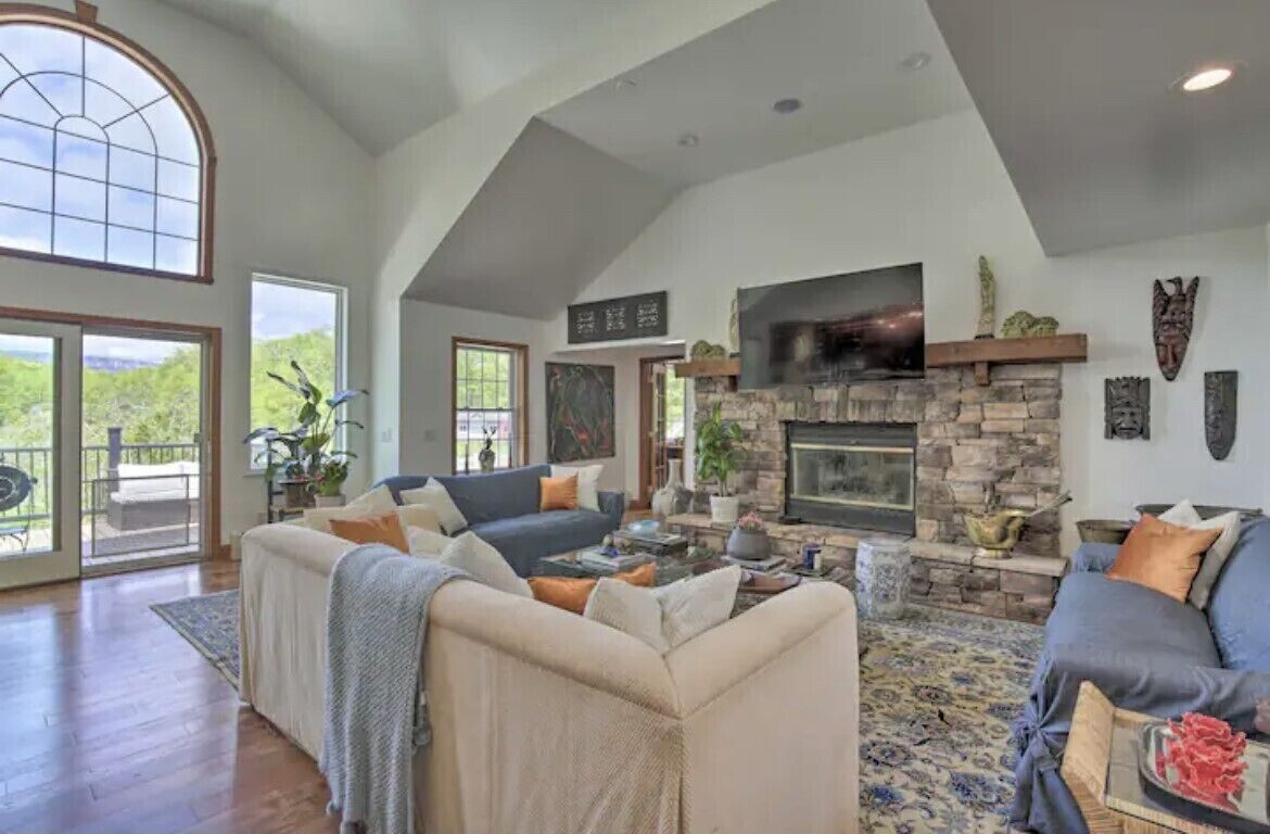 A spacious living room with a stone fireplace, comfortable seating, and large windows.