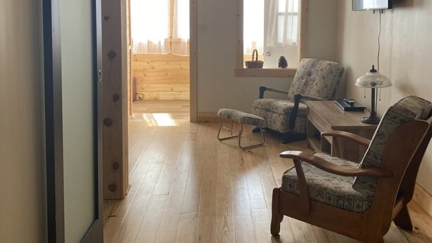 A spacious room with wooden flooring, a large sliding door, two windows with curtains, and a flat-screen TV mounted on the wall.