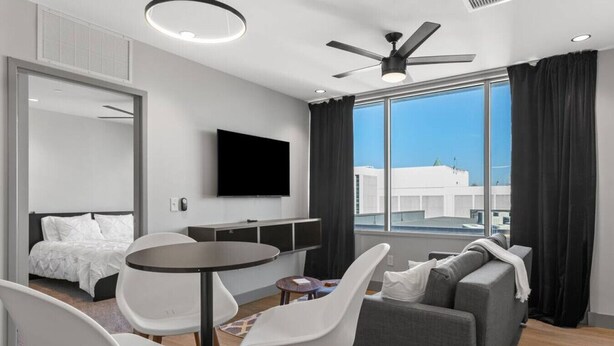 A modern hotel room with a bed, a dining area with a table and chairs, a sofa, a flat-screen TV mounted on the wall, and a large window offering a city view. - 175 LEX - Quiet Loft Near Rupp Arena & Restaurants (Lexington)